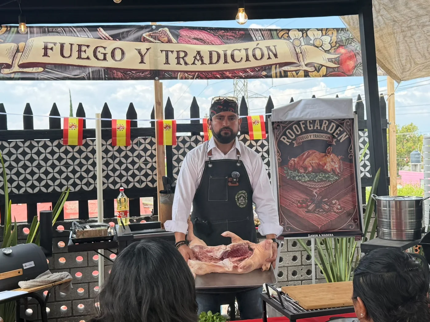 Chef instructor de RoofGarden explicando el corte de un lechón frente al grupo, bajo el letrero Fuego y Tradición